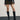 Woman wearing a black minimalist mini skirt that falls mid-thigh, standing confidently. The skirt has a simple, yet elegant design with clean lines and no visible embellishments. She pairs the skirt with a tucked-in white blouse and black pumps for a sophisticated look